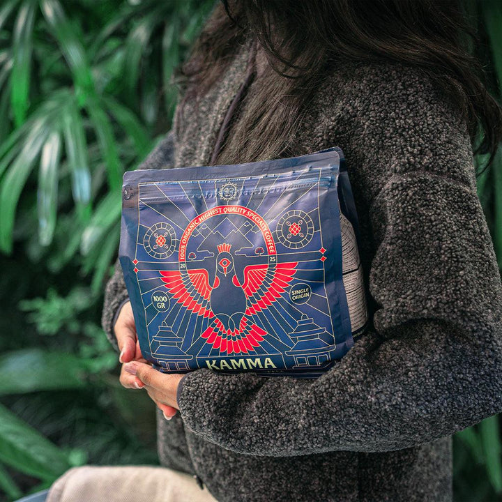 Person holding a blue and red bag with 'KAMMA' branding against a green leafy background